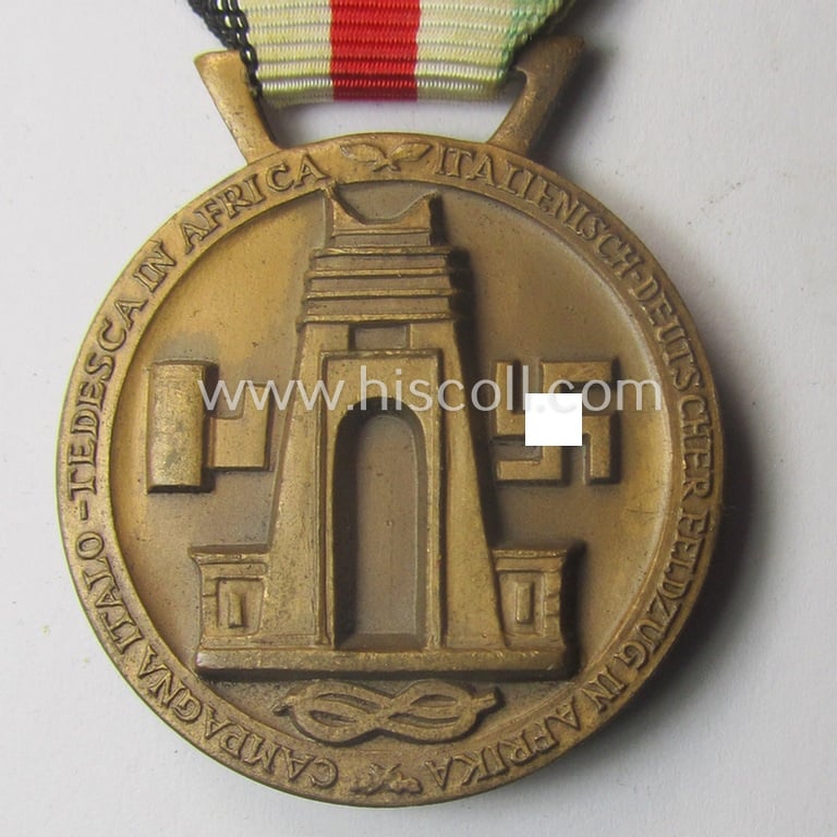 Golden-bronze-coloured- (and I deem 'Buntmetall'-based-) example of a: 'Deutsch-Italienische Feldzugsmedaille' (or: German-Italian campaign-medal) that comes mounted onto its (regular-sized) piece of original (and somewhat faded!) ribbon