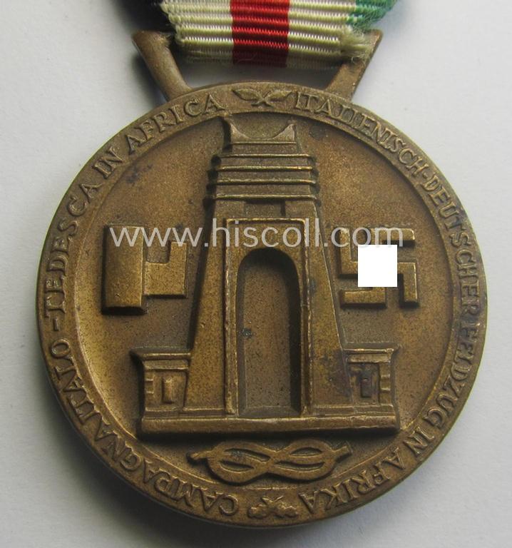 Golden-bronze-coloured- (and I deem 'Buntmetall'-based-) example of a: 'Deutsch-Italienische Feldzugsmedaille' (or: German-Italian campaign-medal) that comes mounted onto its (regular-sized) piece of original (and minimally faded!) ribbon