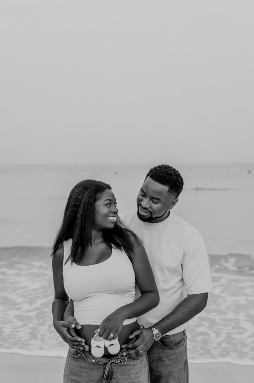 Black and white expecting couple holding tiny white baby shoes in front of her bump at the beach.