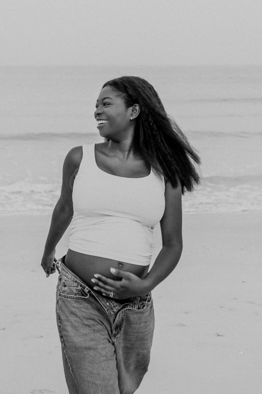 Black and white pregnant woman smiling beside the sea with her hands resting on her bump.