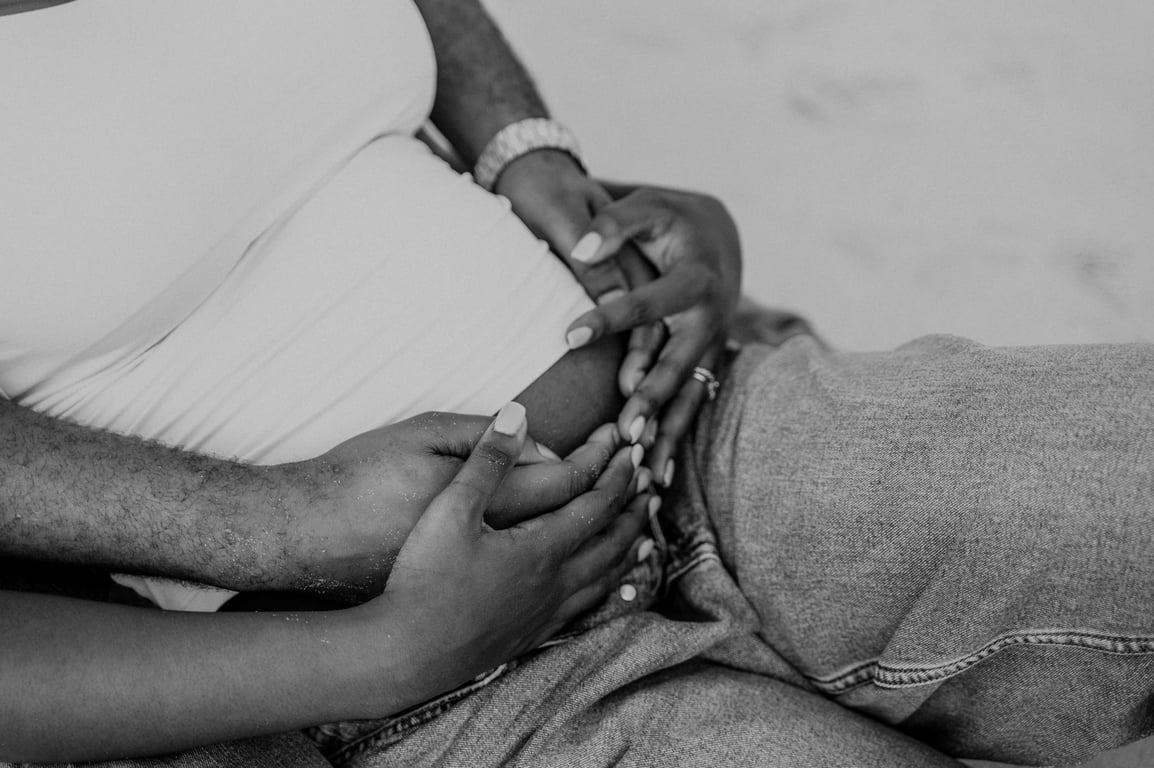 Black and white close-up of the couple’s hands resting together on her bump as they sit on the sand.