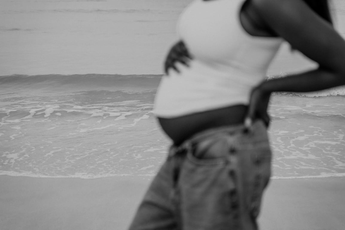 Black and white detail of a pregnant woman’s bump above blue jeans, with her hands at her waist and the waves behind her.