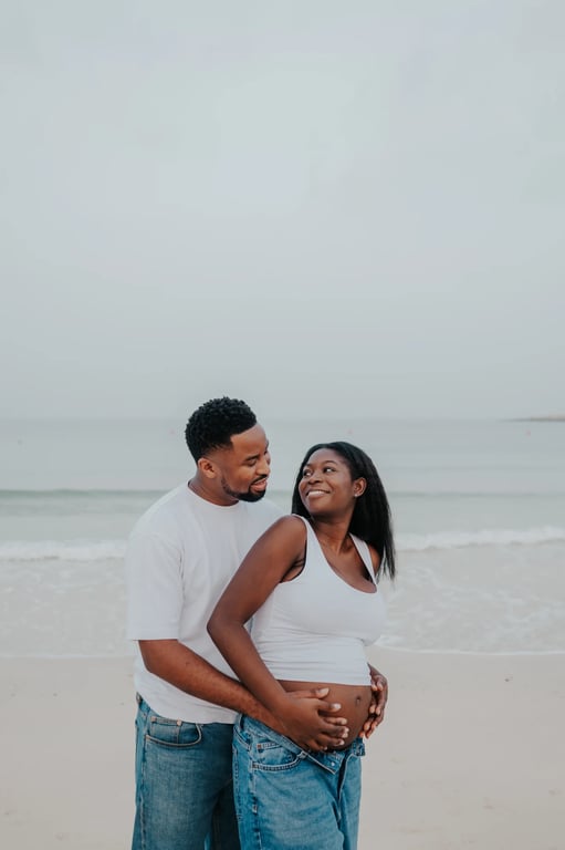 Expecting couple embracing from behind beside the sea, with their hands together on her bump.