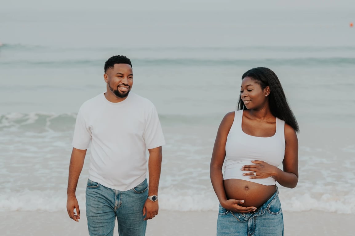 Pregnant woman and her partner walking side by side on the sand, with waves behind them.