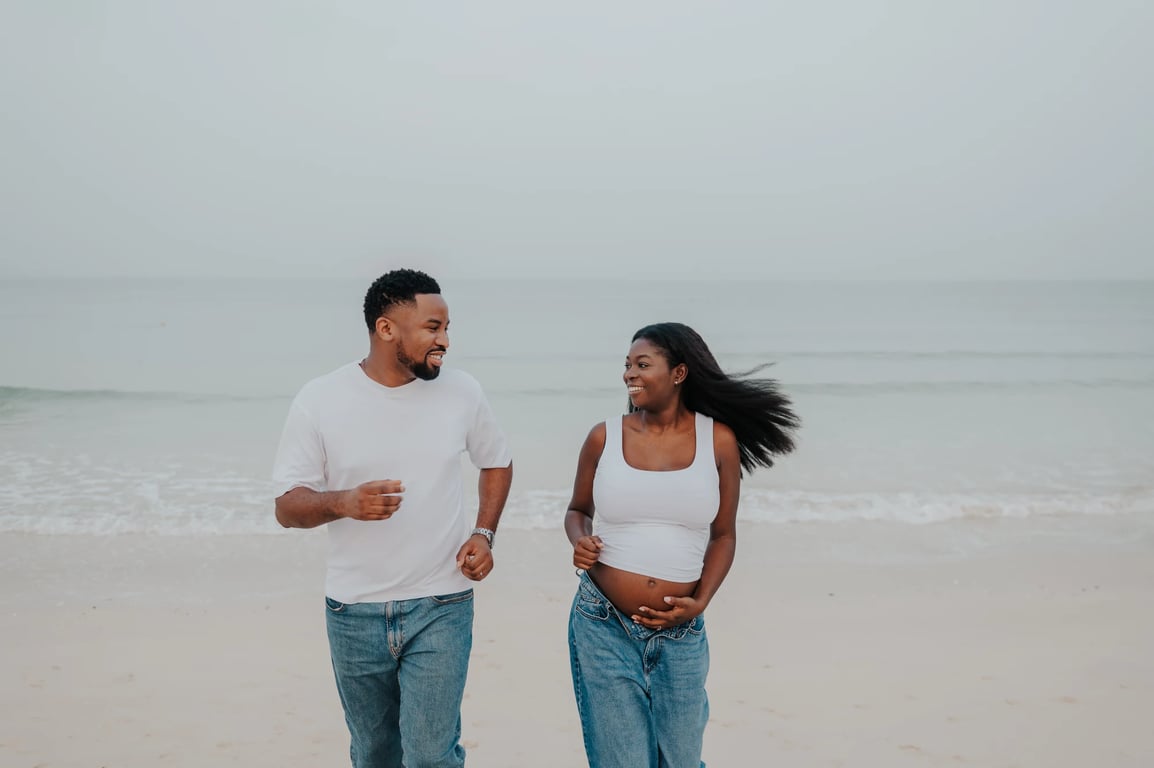 Expecting couple walking together beside the waves in white tops and blue jeans.