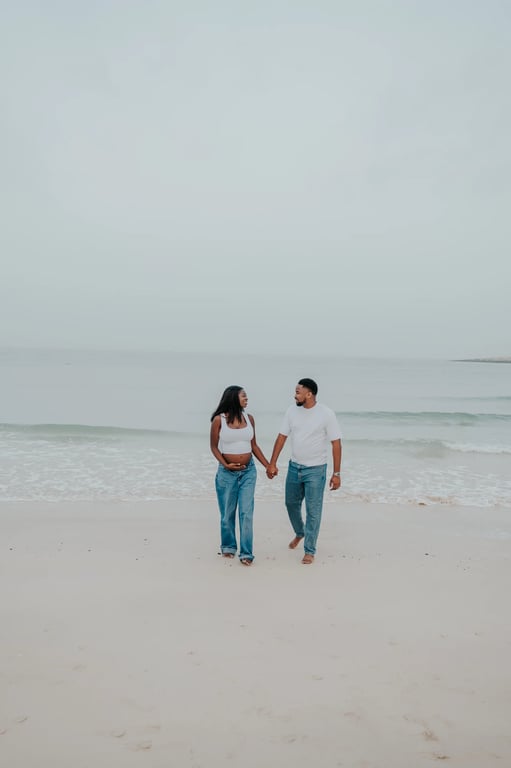 Expecting couple in white tops and blue jeans holding hands on the sand beside the sea in Dubai.