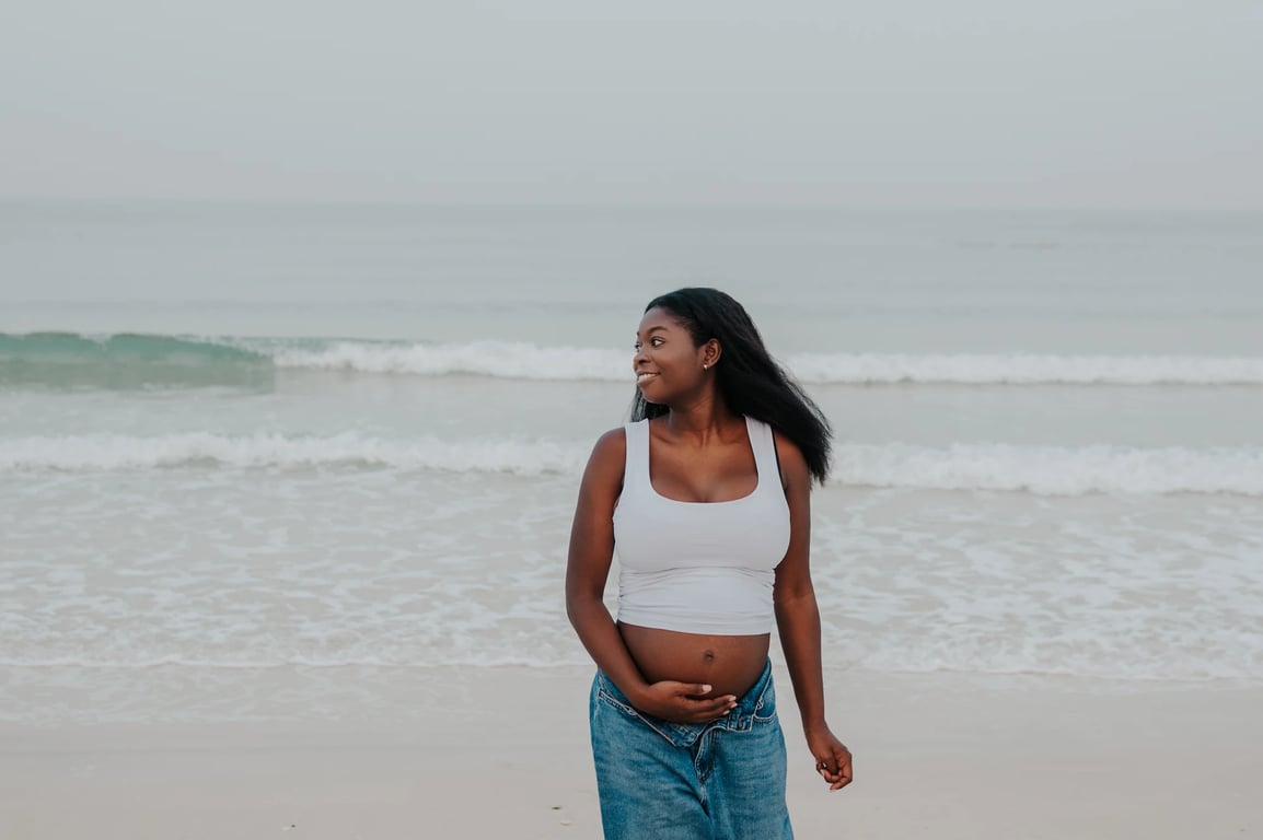Pregnant woman smiling in a white top and blue jeans, with waves behind her.