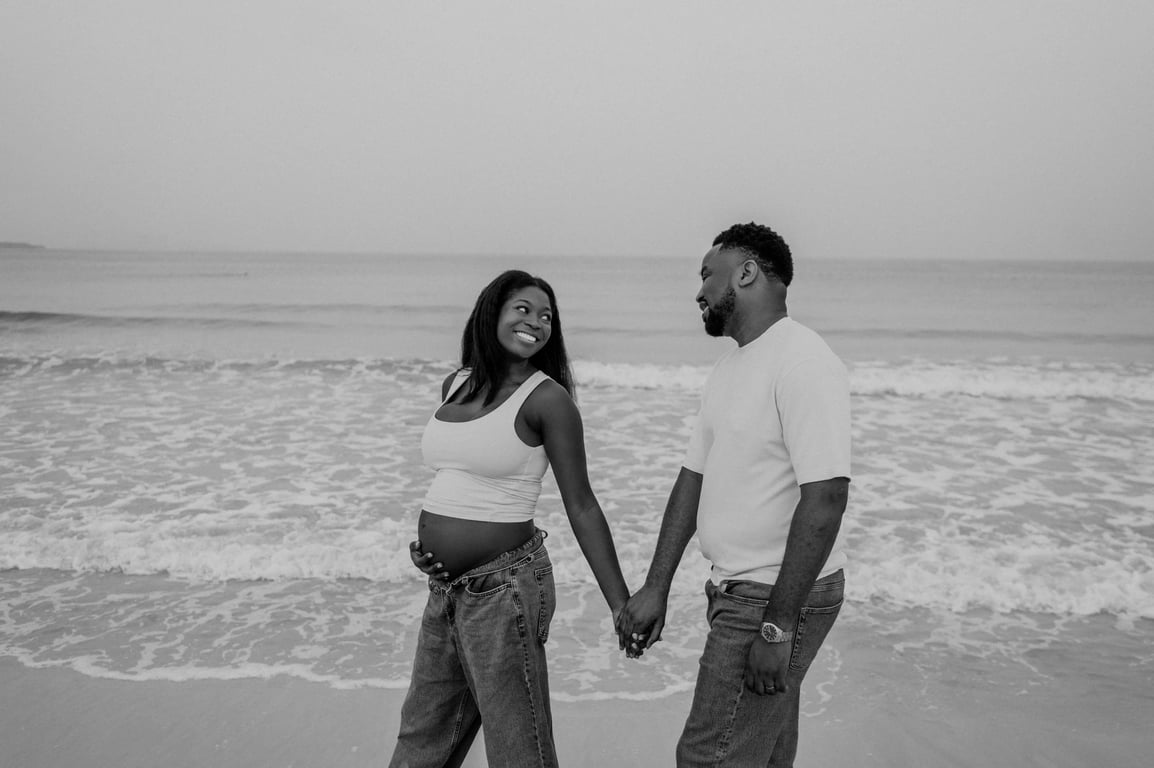 Black and white expecting couple smiling and holding hands in front of the waves during their Dubai beach maternity photoshoot.