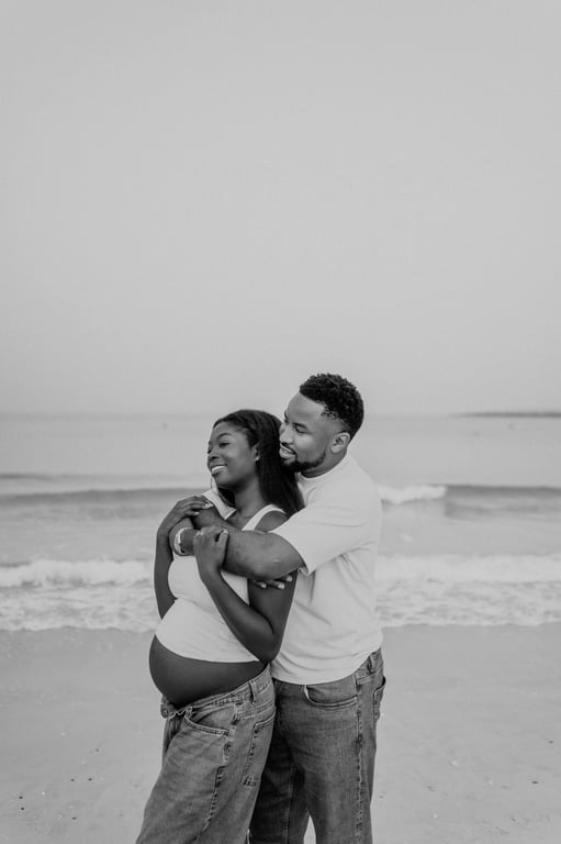 Black and white man embracing a pregnant woman from behind as they stand beside the waves.