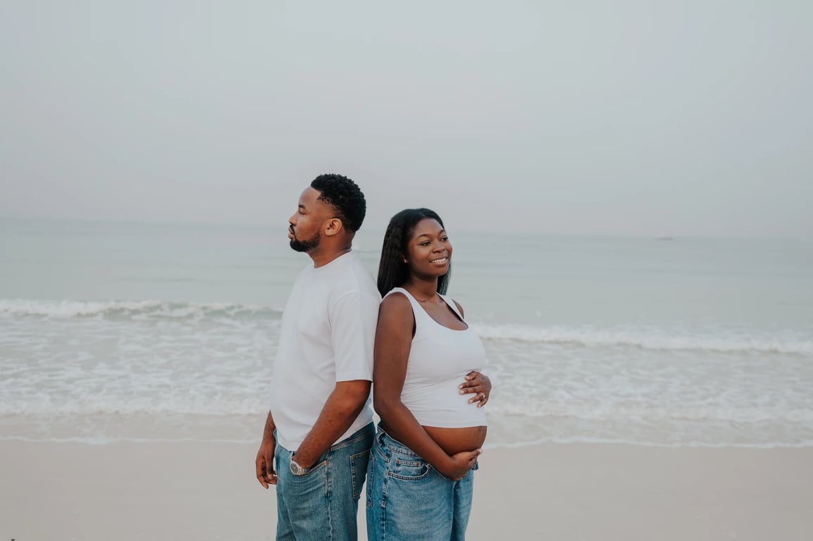 Expecting couple standing back to back beside the sea, with her hands resting on her bump.