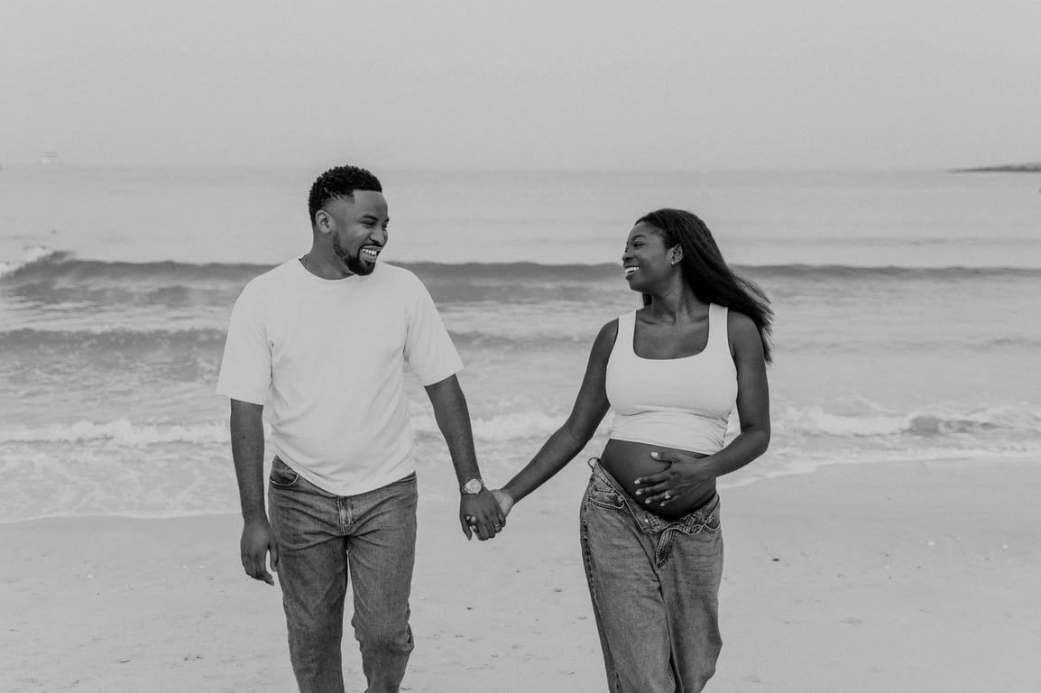 Black and white expecting couple smiling and holding hands in front of the waves during their Dubai beach maternity photoshoot.