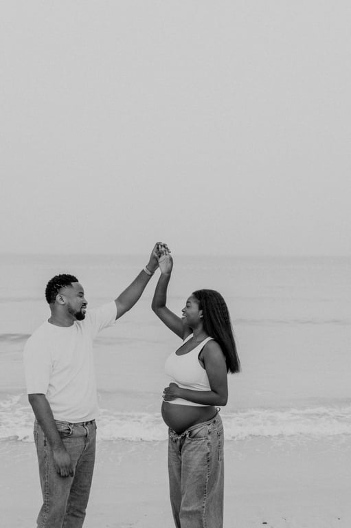Black and white expecting couple lifting their joined hands on the sand beside the sea.