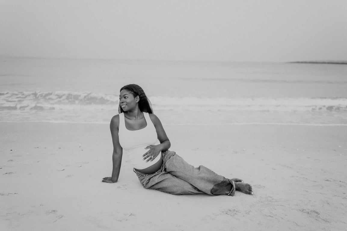 Black and white pregnant woman seated barefoot on pale sand, with one hand resting on her bump and waves behind her.