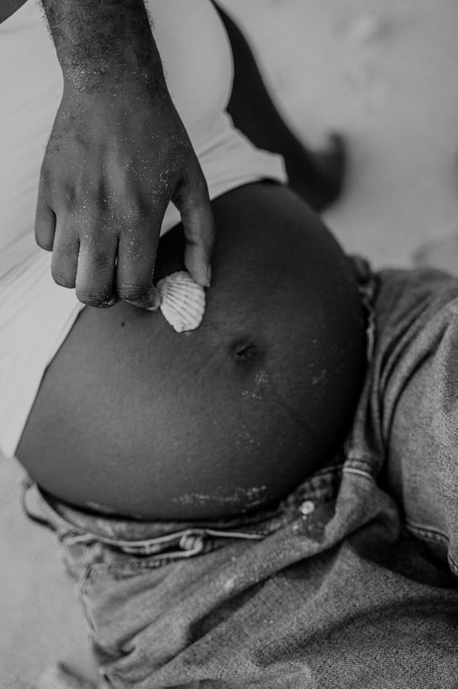 Black and white close-up of a small white shell held against a pregnant woman’s bump above her blue jeans.