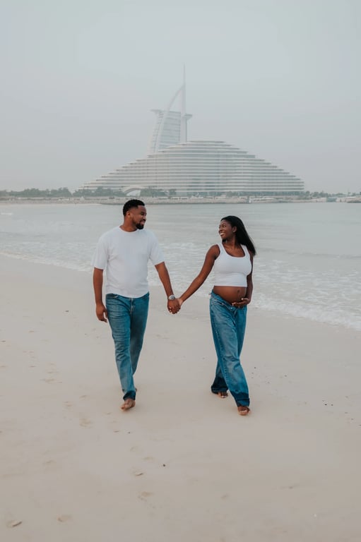 Expecting couple holding hands and walking on the beach, with Burj Al Arab in the background.