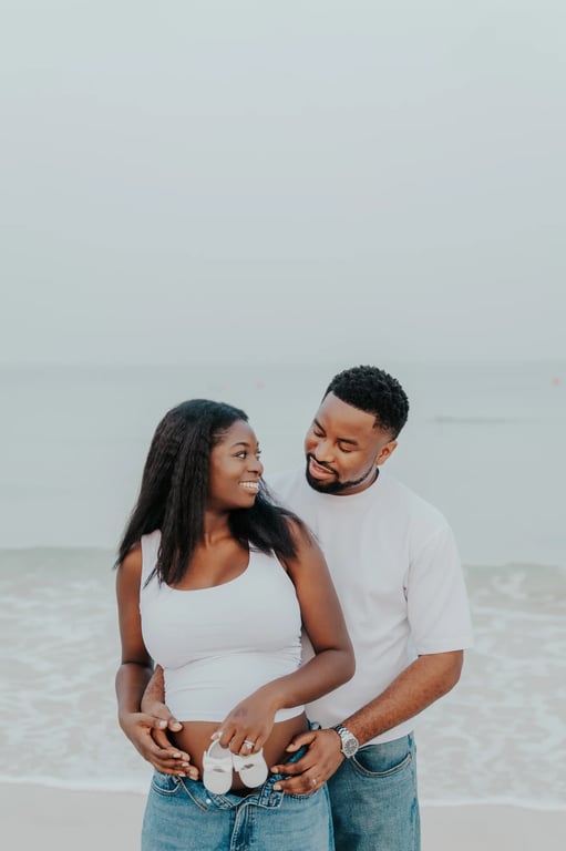 Expecting couple holding tiny white baby shoes in front of her bump at the beach.