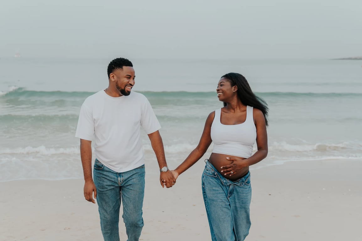 Expecting couple smiling and holding hands in front of the waves during their Dubai beach maternity photoshoot.