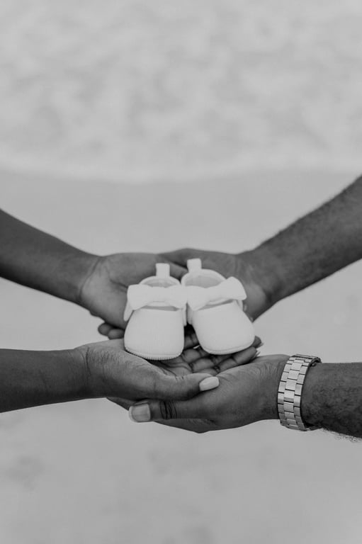 Black and white close-up of the couple’s hands holding a pair of tiny white baby shoes beside the sea.