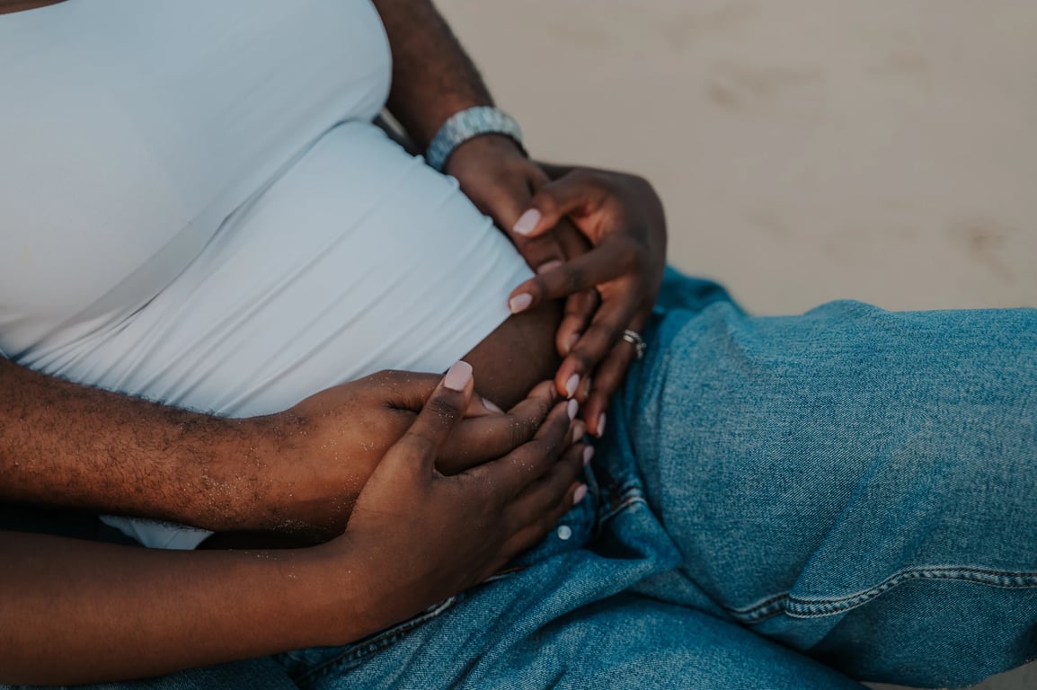 Close-up of the couple’s hands resting together on her bump as they sit on the sand.