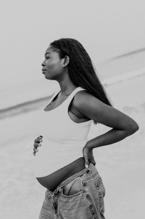 Black and white side-on maternity portrait of a woman in a white top and blue jeans beside the sea.
