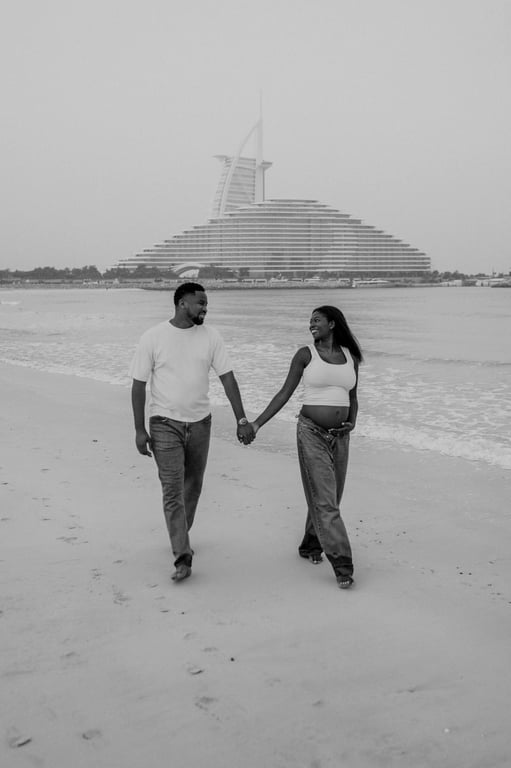 Black and white expecting couple holding hands and walking on the beach, with Burj Al Arab in the background.