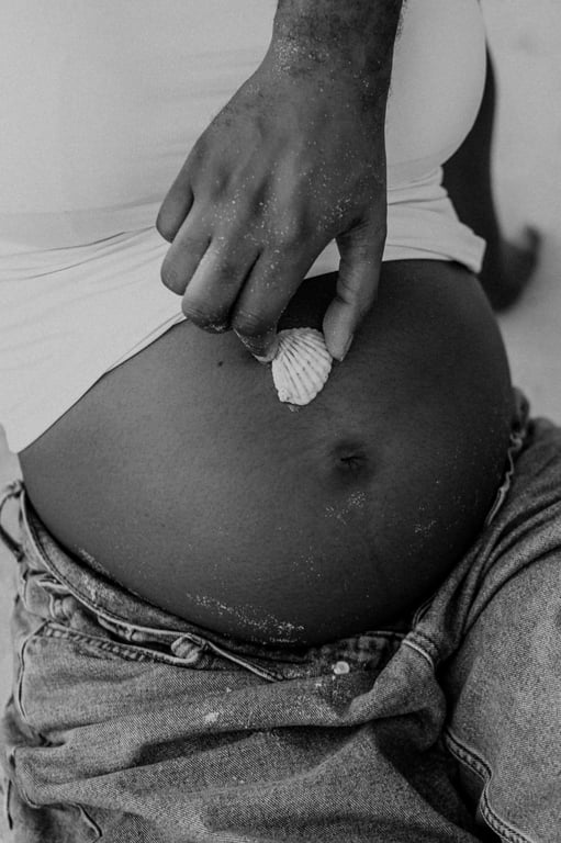 Black and white close-up of a small white shell held against a pregnant woman’s bump above her blue jeans.