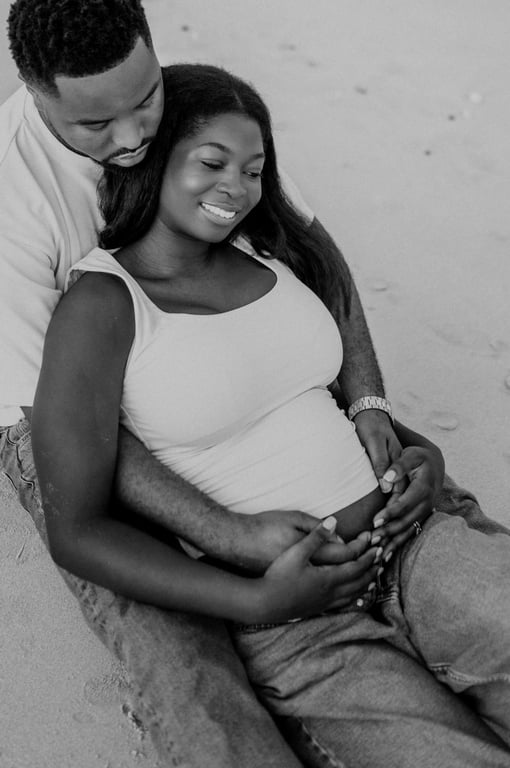 Black and white expecting couple seated together on the sand, with their hands resting on her bump.