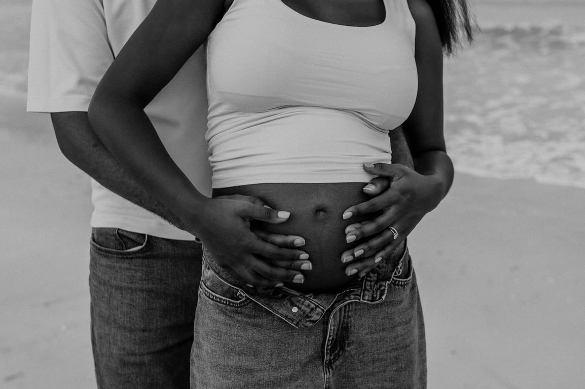 Black and white close-up of the couple’s hands holding her bump, with white tops and blue jeans.