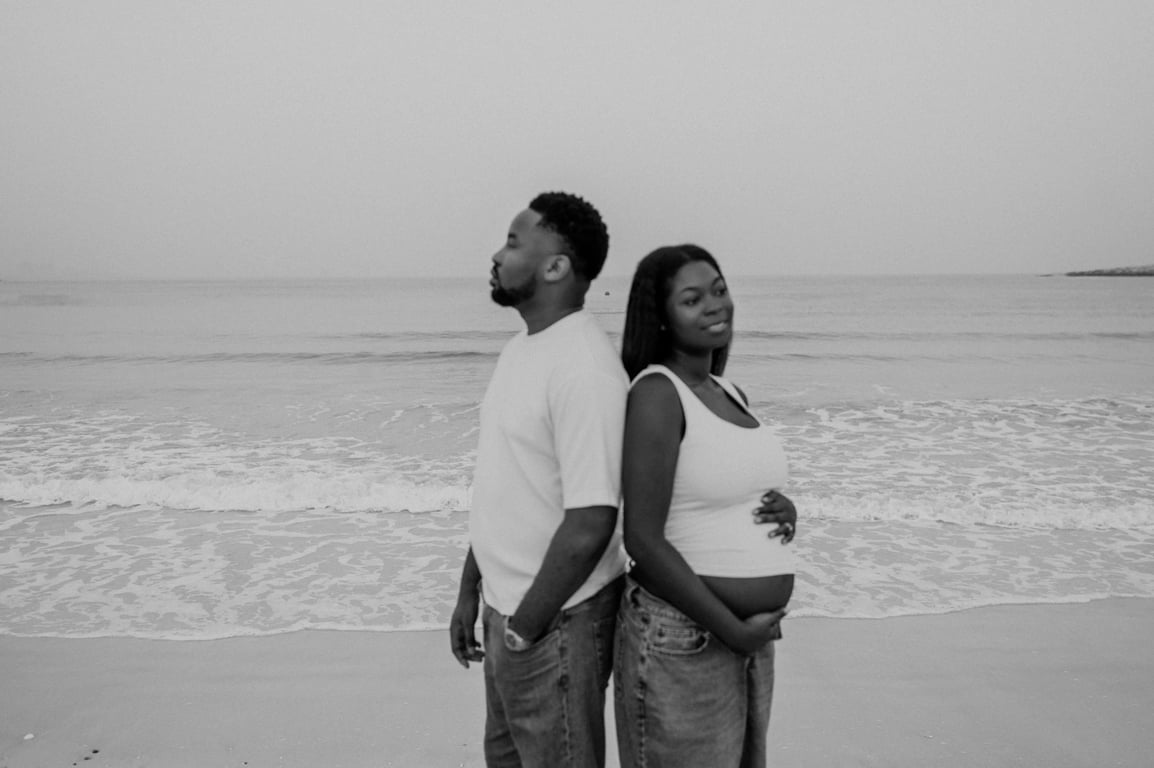 Black and white expecting couple standing back to back beside the sea, with her hands resting on her bump.