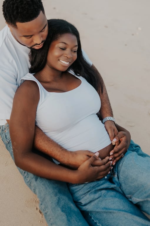 Expecting couple seated together on the sand, with their hands resting on her bump.