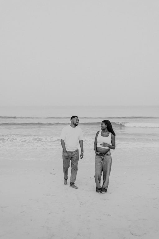 Black and white pregnant woman and her partner walking side by side on the sand, with waves behind them.