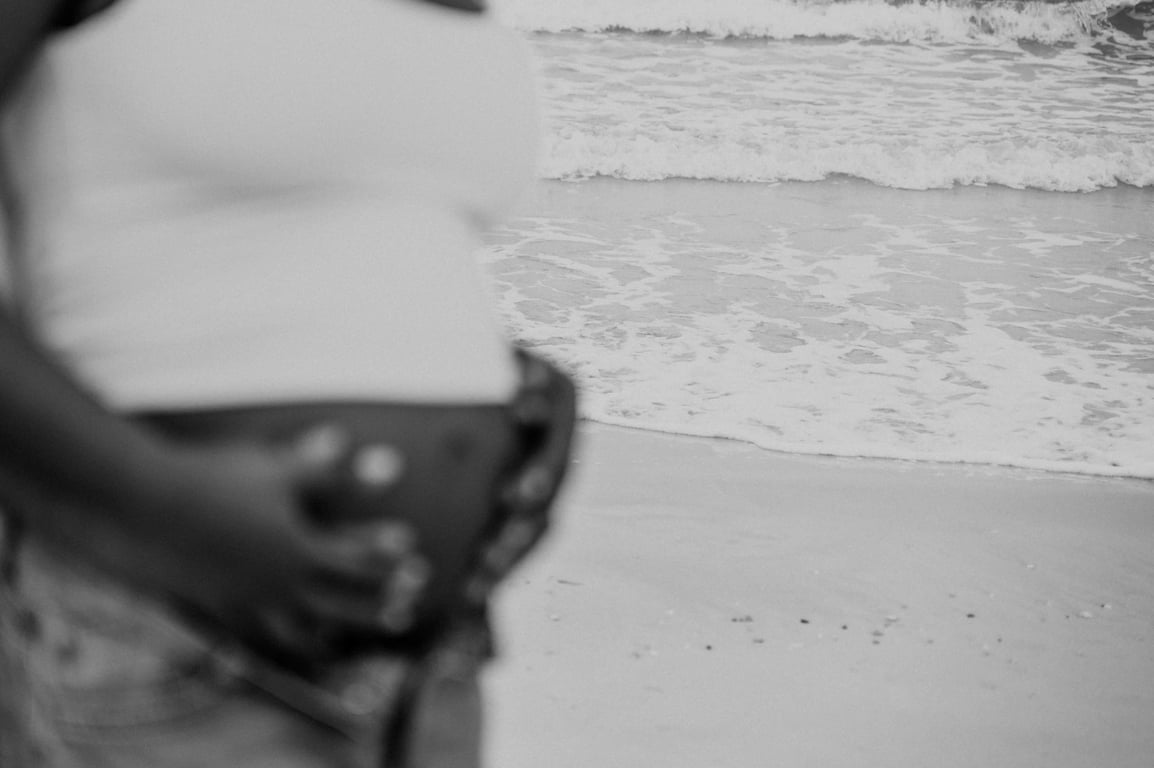 Black and white soft-focus detail of a pregnant woman holding her bump beside the waves.