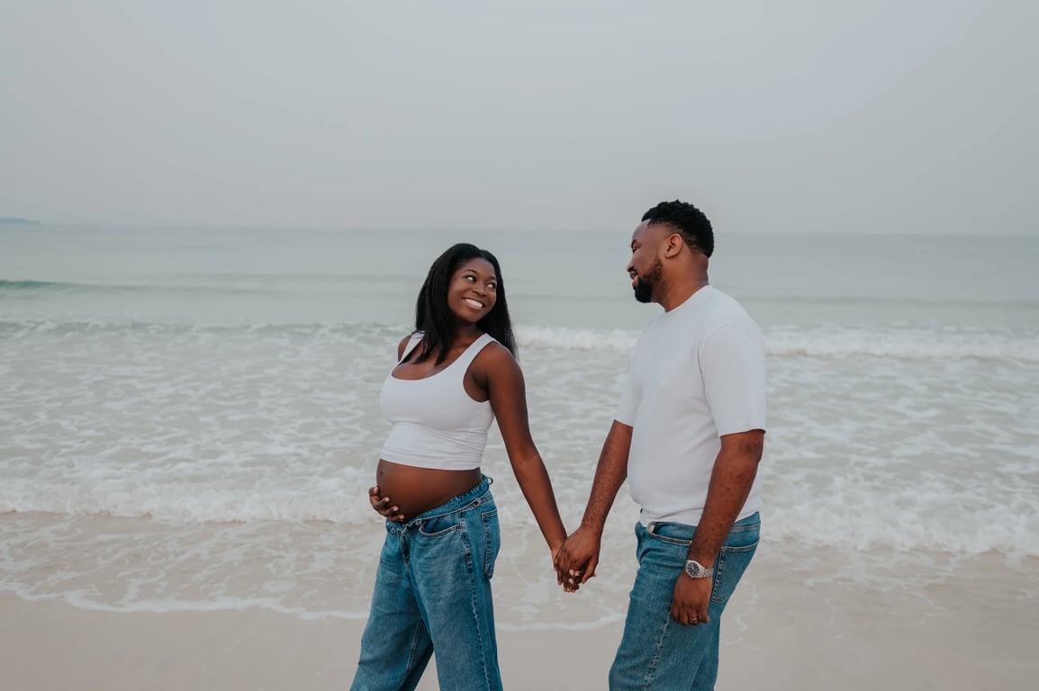 Expecting couple smiling and holding hands in front of the waves during their Dubai beach maternity photoshoot.