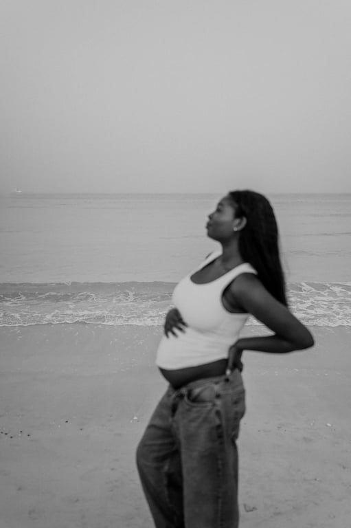 Black and white side-on maternity portrait of a woman in a white top and blue jeans beside the sea.