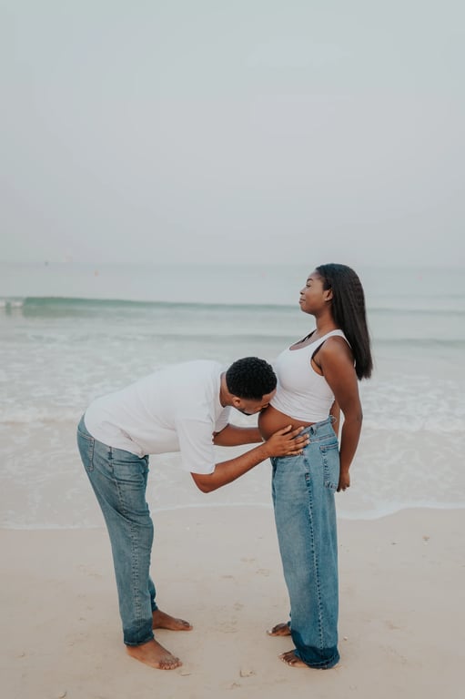 Man bending to kiss a pregnant woman’s bump beside the waves.