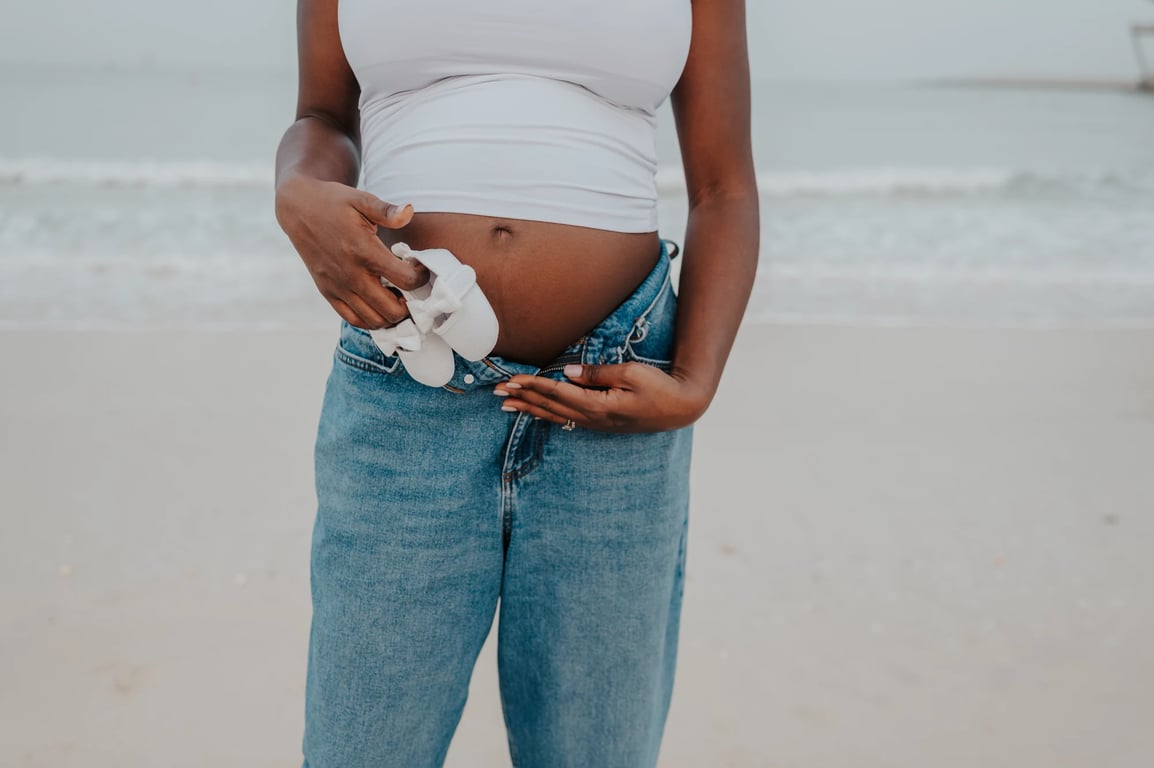 Pregnant woman holding tiny white baby shoes beside her bump at the beach.