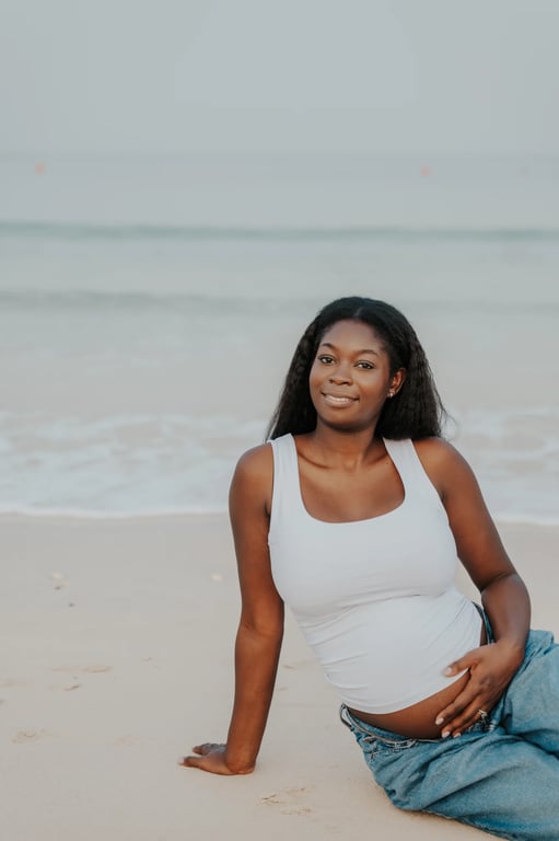 Pregnant woman seated barefoot on pale sand, with one hand resting on her bump and waves behind her.