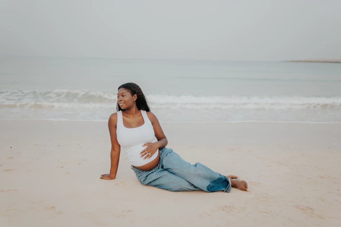 Pregnant woman seated barefoot on pale sand, with one hand resting on her bump and waves behind her.