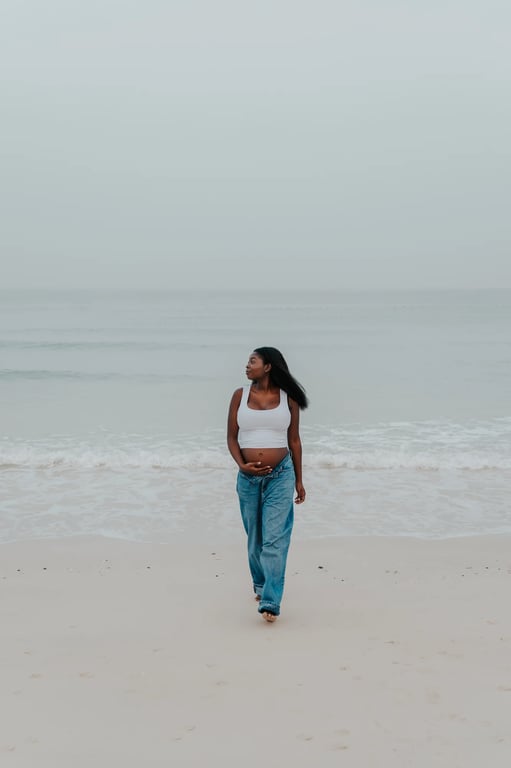Pregnant woman in a white top and blue jeans walking barefoot along the Dubai shoreline.