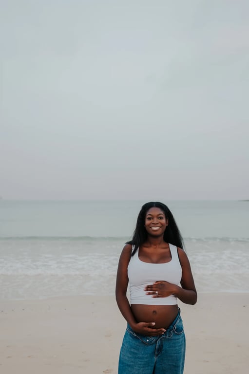 Pregnant woman smiling in a white top and blue jeans, with waves behind her.