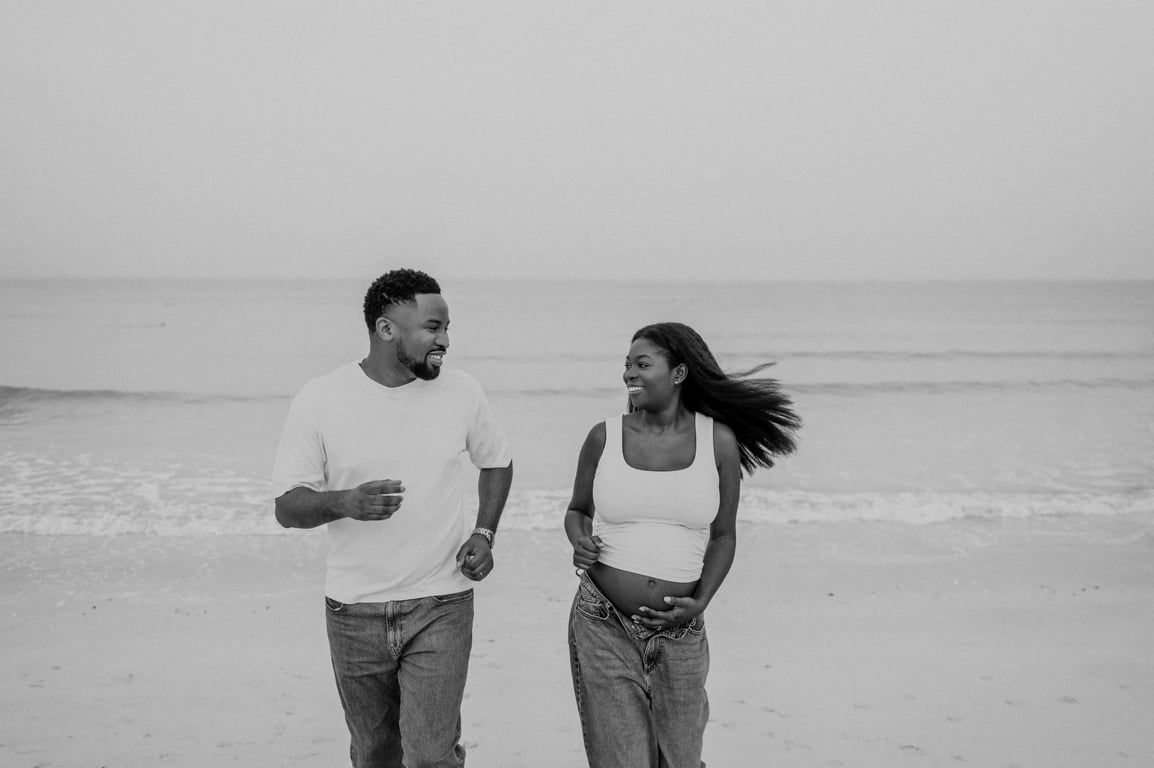 Black and white expecting couple walking together beside the waves in white tops and blue jeans.