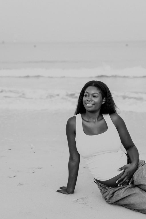 Black and white pregnant woman seated barefoot on pale sand, with one hand resting on her bump and waves behind her.