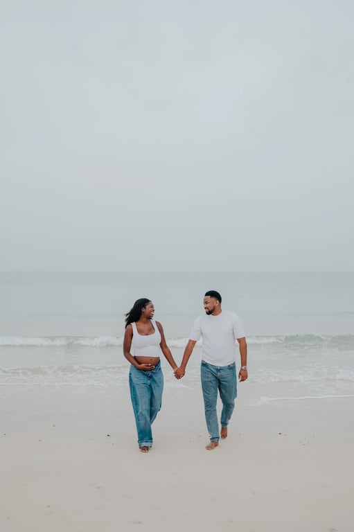 Expecting couple in white tops and blue jeans holding hands on the sand beside the sea in Dubai.