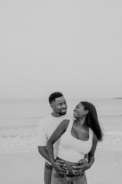 Black and white expecting couple embracing from behind beside the sea, with their hands together on her bump.