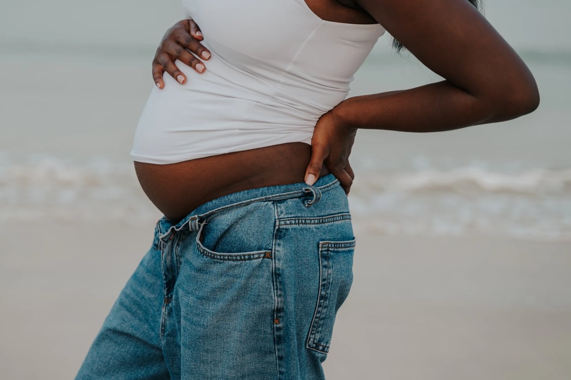 Detail of a pregnant woman’s bump above blue jeans, with her hands at her waist and the waves behind her.