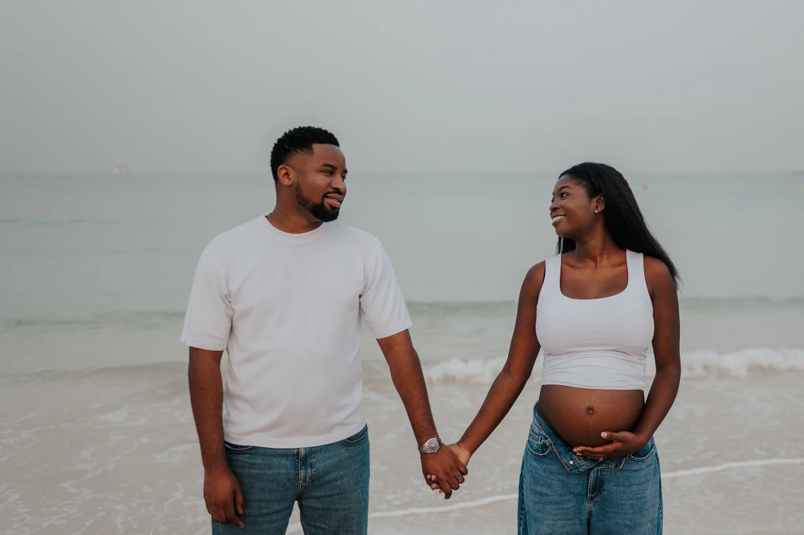 Expecting couple smiling and holding hands in front of the waves during their Dubai beach maternity photoshoot.