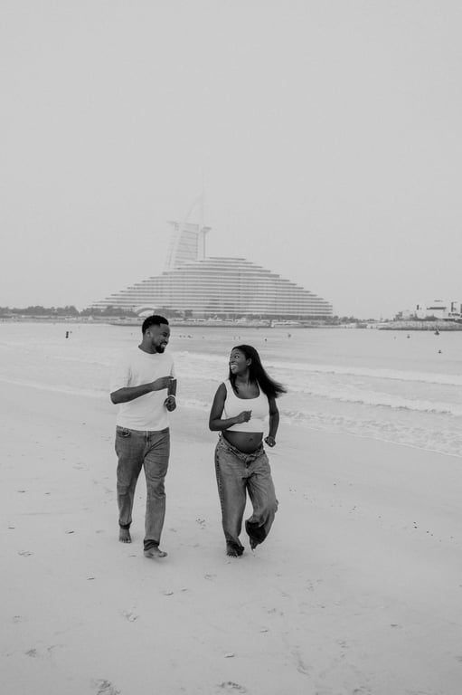 Black and white expecting couple walking together beside the waves in white tops and blue jeans.