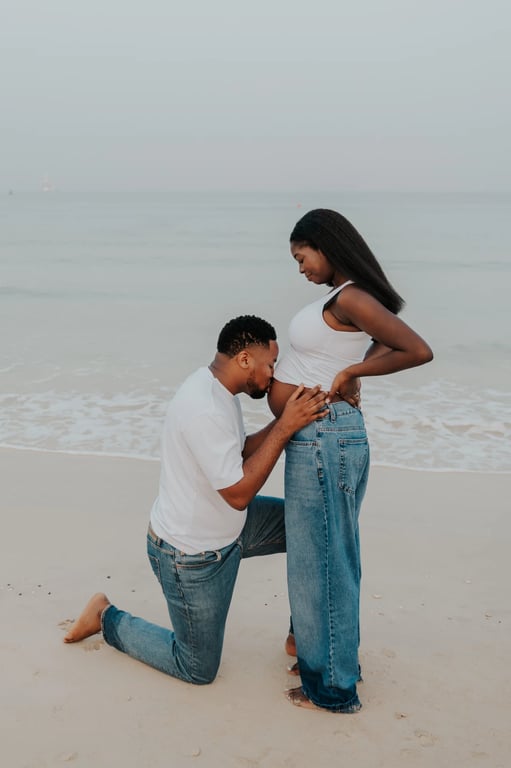 Man kneeling to kiss a pregnant woman’s bump on the sand beside the sea.