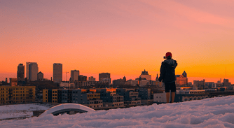 Person standing on snow-covered rooftop at sunset with city skyline in background