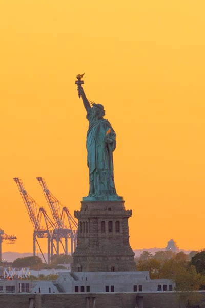 The Statue of Liberty and distant city lights under a deep red sunset sky